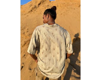 Op de afbeelding: Een persoon draagt een beige shirt met korte mouwen met een bruin geometrisch patroon. Het shirt heeft een knoopsluiting aan de voorkant en een ontspannen pasvorm.