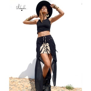 Puede incluir: Un top corto negro y una falda negra única con un panel estampado beige y negro. La falda tiene paneles laterales largos y fluidos y está adornada con tachuelas metálicas. Un sombrero negro y joyas completan el look. El texto "Melmakka..." está en la esquina superior izquierda.