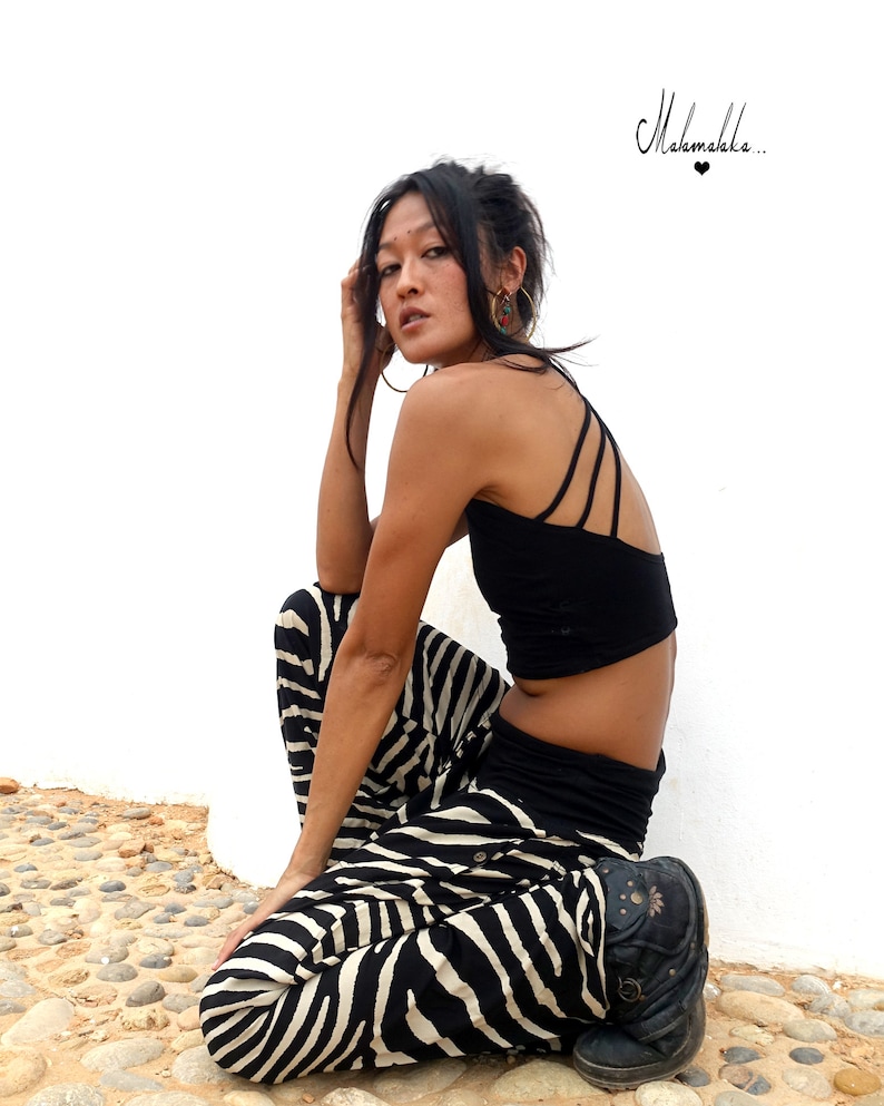 May include: A woman wearing a black crop top and zebra print trousers sits on a bed of small stones. The text "Makamaka..." is written in black on a white background above her head.