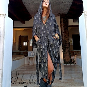 May include: A long, hooded black dress with a fan-like gold pattern. The dress has a high slit, pockets, and a button-down front. The model is wearing black boots and gold bracelets. The dress is tied at the waist with matching fabric.