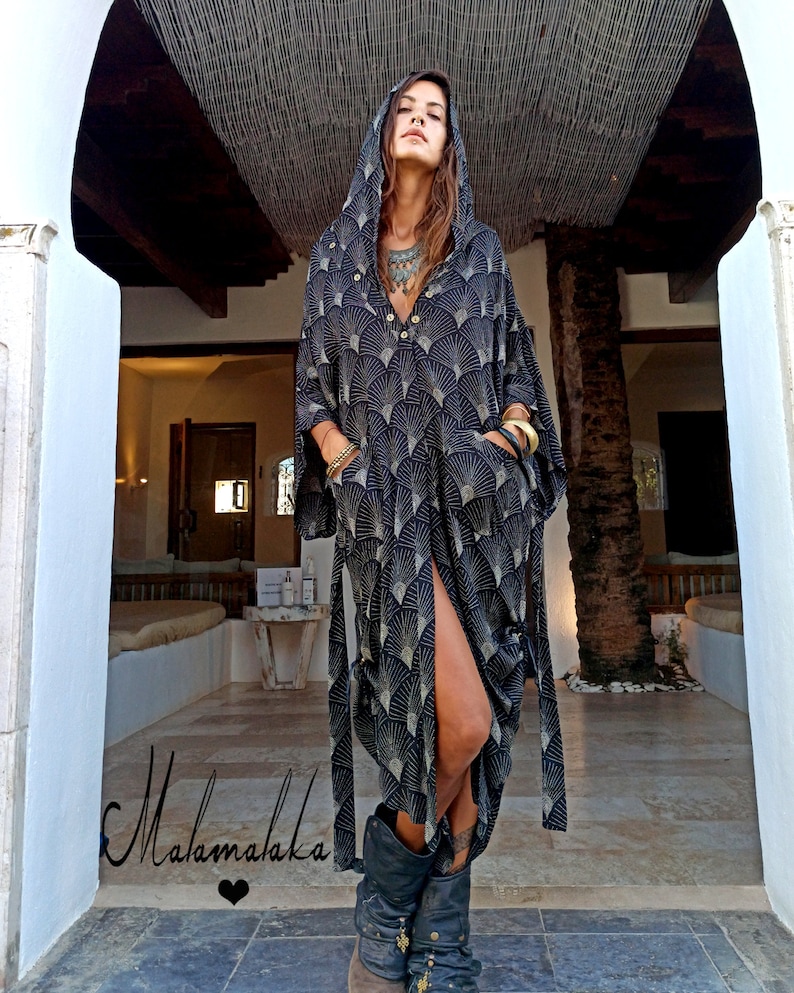 May include: A long, hooded black dress with a fan-like gold pattern. The dress has a high slit, pockets, and a button-down front. The model is wearing black boots and gold bracelets. The dress is tied at the waist with matching fabric.
