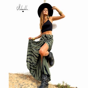 Puede incluir: Un top corto negro y una falda larga verde oliva con rayas de cebra negras. La falda tiene una abertura alta. La modelo lleva un sombrero y botas negras. Pulseras y pendientes dorados completan el look. El texto "Mahmekka..." está en la esquina superior izquierda.