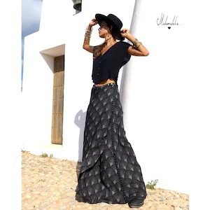 May include: A black wrap skirt with a fan-like pattern, paired with a black top and hat. The skirt is floor-length and has a unique design. Gold bracelets adorn the arms. The word "Mahamakka" is written on the wall.