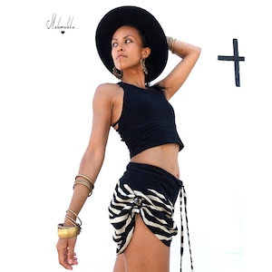 Puede incluir: Top negro y shorts negros con estampado de rayas de cebra blancas. La modelo lleva un sombrero negro y pulseras doradas. La imagen incluye el texto "Mahmekka..." en cursiva.