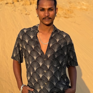 Op de afbeelding: Zwart overhemd met knopen en een zich herhalend waaiervormig patroon in beige. Het shirt met korte mouwen wordt gedragen met een kaki broek en een zilveren ketting en armband. De persoon heeft zijn handen in zijn zakken.