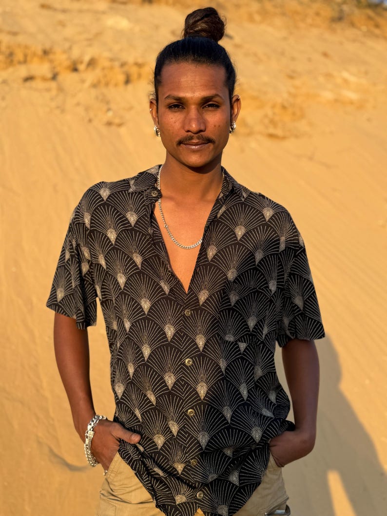Op de afbeelding: Zwart overhemd met knopen en een zich herhalend waaiervormig patroon in beige. Het shirt met korte mouwen wordt gedragen met een kaki broek en een zilveren ketting en armband. De persoon heeft zijn handen in zijn zakken.