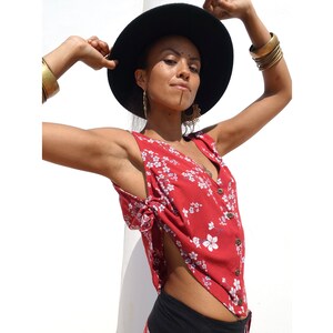 Puede incluir: Una blusa roja sin mangas con estampado floral blanco y cierre de botones. La blusa tiene un detalle anudado en el lateral y se lleva con un sombrero negro.