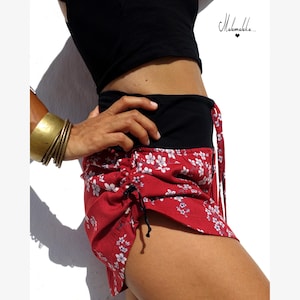 Puede incluir: Shorts rojos con estampado floral blanco y detalles de cordón negro. Los shorts tienen una cintura negra y se combinan con una parte superior negra. Se ve un brazalete dorado y varias pulseras doradas en el brazo.