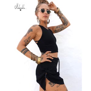 Puede incluir: Conjunto de top y falda negros. El top es sin mangas. La falda tiene una abertura lateral y detalles de cordón. La imagen también incluye múltiples pulseras doradas y gafas de sol en forma de corazón. El texto "Mahamala" está en la esquina superior izquierda.