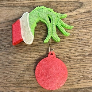 May include: A green and red Grinch hand ornament with a red glitter ornament hanging from it.