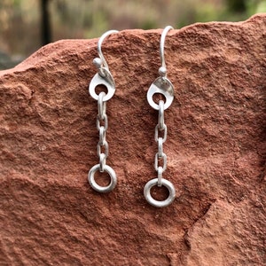 May include: A pair of silver dangle earrings with a chain and a small circle at the bottom. The earrings are hanging from a rock.
