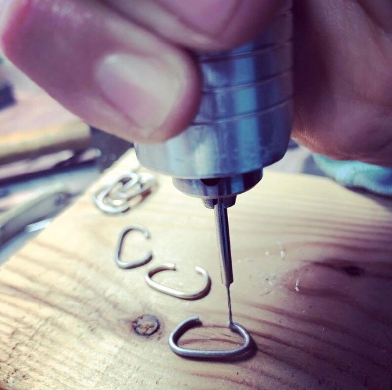 May include: A close-up of a hand using a small drill to create holes in a series of metal rings. The rings are arranged in a row on a wooden surface.
