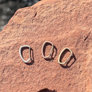 May include: Three metal carabiners in different finishes: silver, gold, and brass. The carabiners are arranged in a row on a red rock surface.