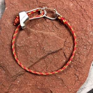 May include: A red and yellow rope bracelet with a silver metal clasp. The bracelet is lying on a red, textured surface.