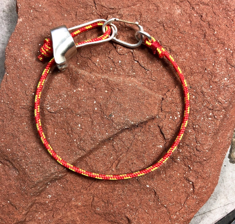 May include: A red and yellow rope bracelet with a silver metal clasp. The bracelet is lying on a red, textured surface.