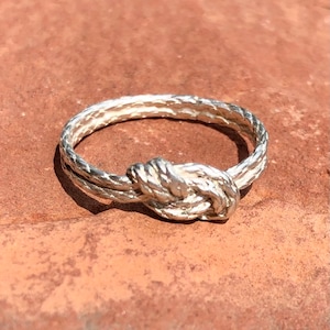 May include: A silver ring with a knotted design. The ring is made of two strands of twisted metal that are tied together in a knot.