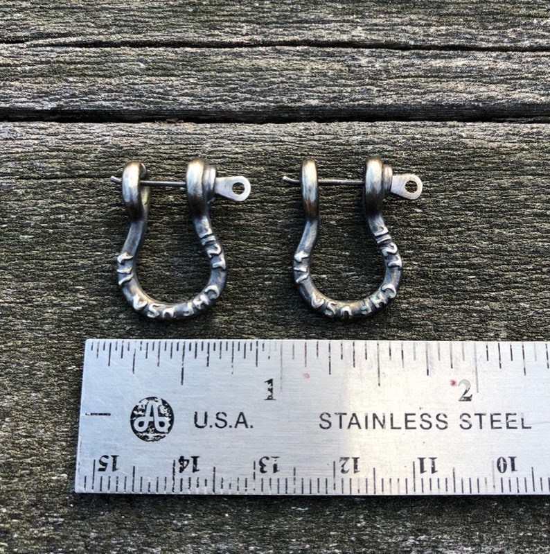 May include: Two silver-toned metal earrings in the shape of a U with a small loop at the top. The earrings are engraved with the words "USA" and "CHIF" on the sides. The earrings are measured against a ruler.