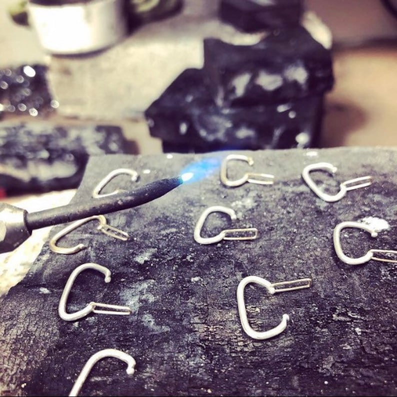 May include: A close-up of a metalworking process, showing a torch heating a small, silver heart-shaped piece of metal. Several other heart-shaped pieces are scattered on a black surface.