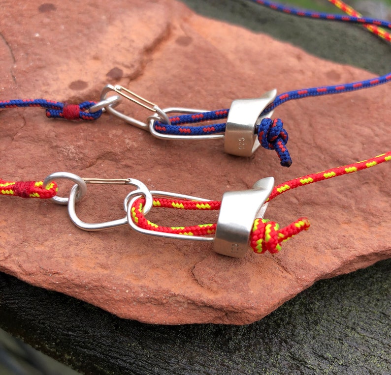 May include: Two silver climbing carabiners with red and yellow ropes attached. The carabiners are on a brown rock surface.
