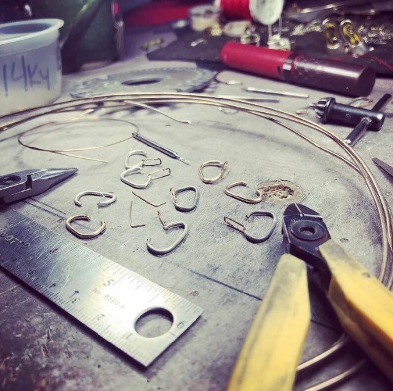 May include: A close-up of a workbench with various jewelry making supplies, including wire, pliers, and small metal hoops. The workbench is made of metal and has a gray surface. There are also several tools and other supplies scattered around the workbench.