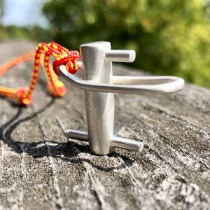 May include: A silver metal pendant shaped like a hook with a red and yellow cord attached. The pendant is on a wooden surface.