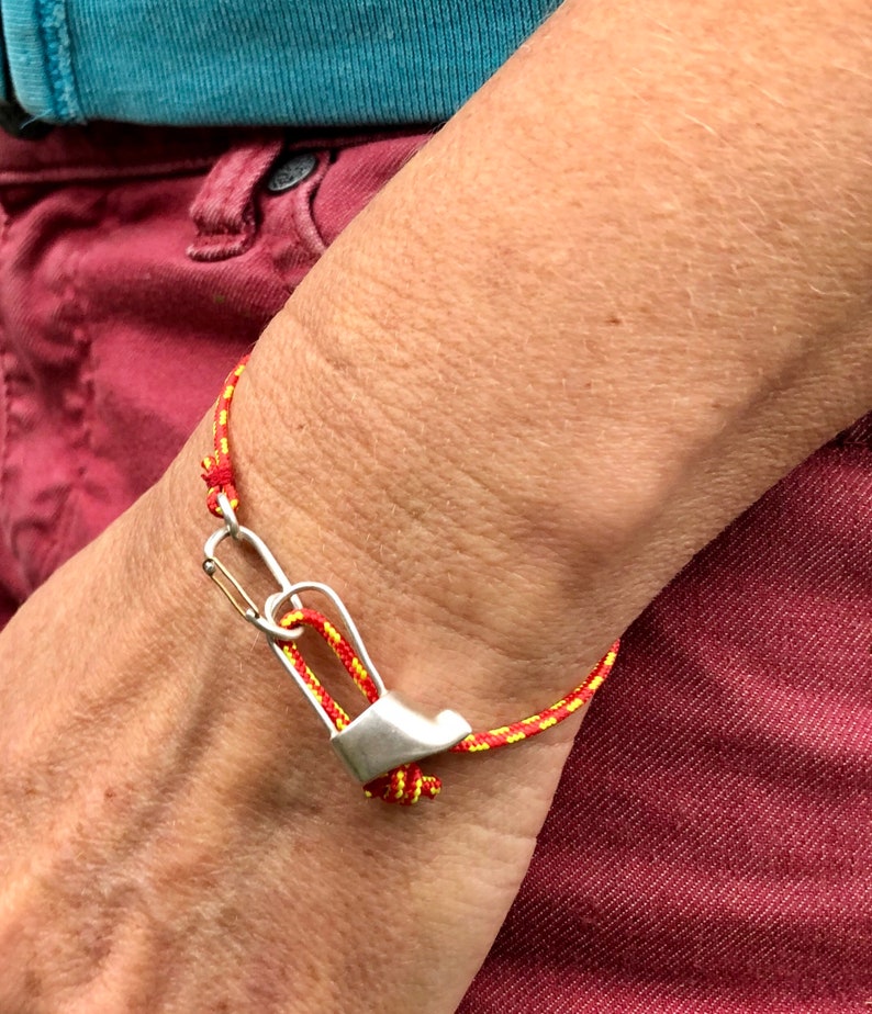 May include: A red and yellow braided bracelet with a silver carabiner clasp. The bracelet is worn on a person's wrist.