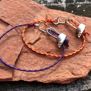 May include: Two bracelets made with red, yellow, and blue cord and silver metal clasps. The bracelets are tied together with a knot.