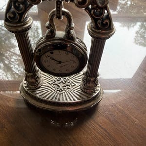 Può includere: Un orologio da tavolo in metallo color argento sospeso all'interno di una cornice ad arco con due colonne scanalate e una base decorativa. Il quadrante è bianco con numeri neri e un piccolo simbolo a cuore alle ore dodici.