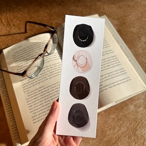 May include: A bookmark featuring four watercolor illustrations of cowboy hats in different colors and styles. The hats are arranged vertically on a white background.