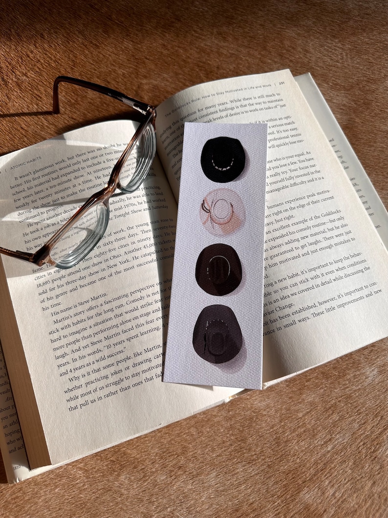 May include: A close-up of a bookmark with a watercolor illustration of three black cowboy hats. The bookmark is on top of an open book with the text "How to Stay Motivated in Life and Work" visible.