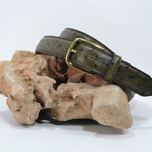 Peut inclure: Une ceinture en cuir d'autruche verte avec une boucle en laiton. La ceinture est enroulée et repose sur un rocher.