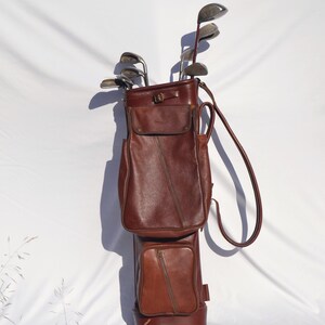 May include: A brown leather golf bag with a zipped pocket and a strap. The bag is filled with golf clubs.