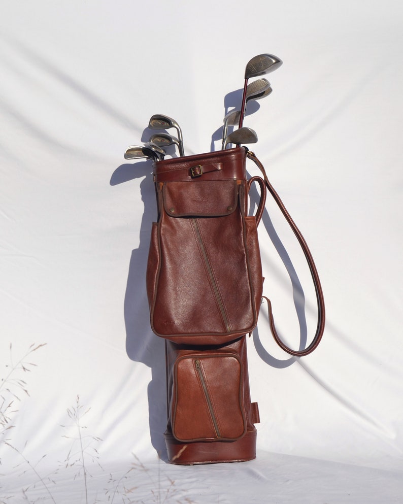 May include: A brown leather golf bag with a zipped pocket and a strap. The bag is filled with golf clubs.