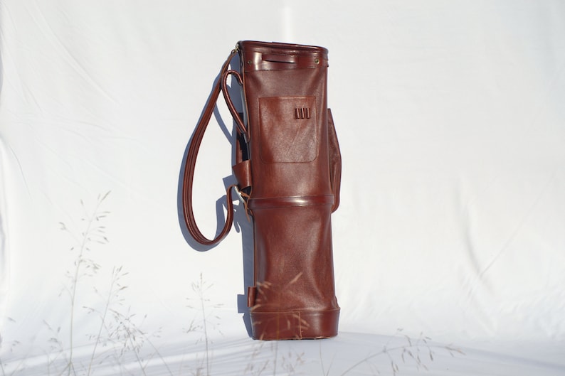 May include: A brown leather golf bag with a single strap and a small pocket on the front.