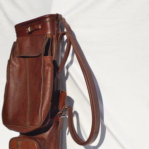 May include: A brown leather golf bag with a long strap and multiple pockets.