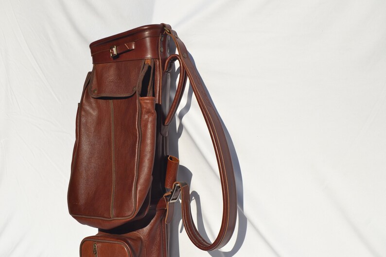 May include: A brown leather golf bag with a long strap and multiple pockets.