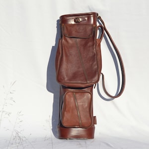 May include: A brown leather golf bag with a single strap and multiple pockets.