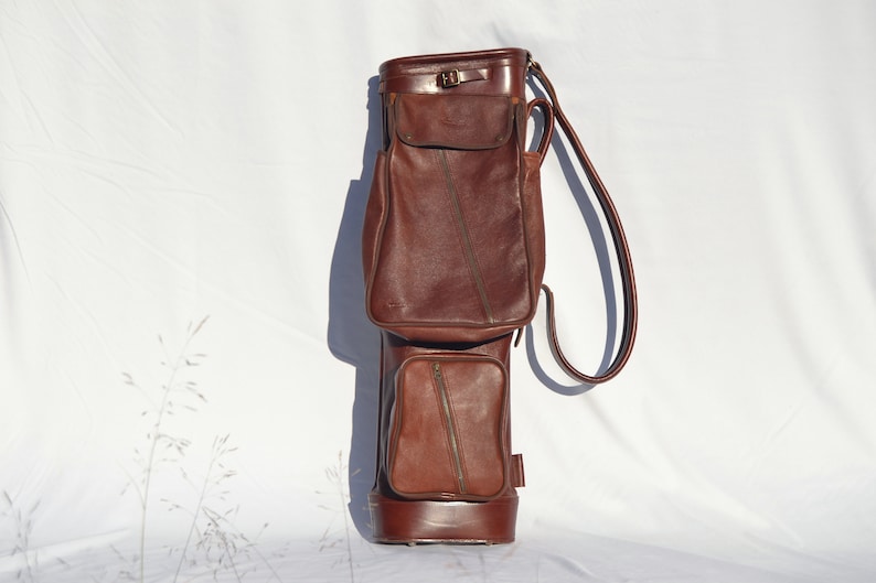 May include: A brown leather golf bag with a single strap and multiple pockets.