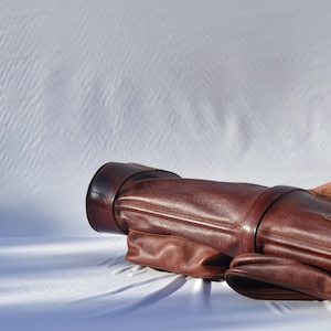 May include: A brown leather golf bag with a strap and buckle. The bag is lying on a white surface.