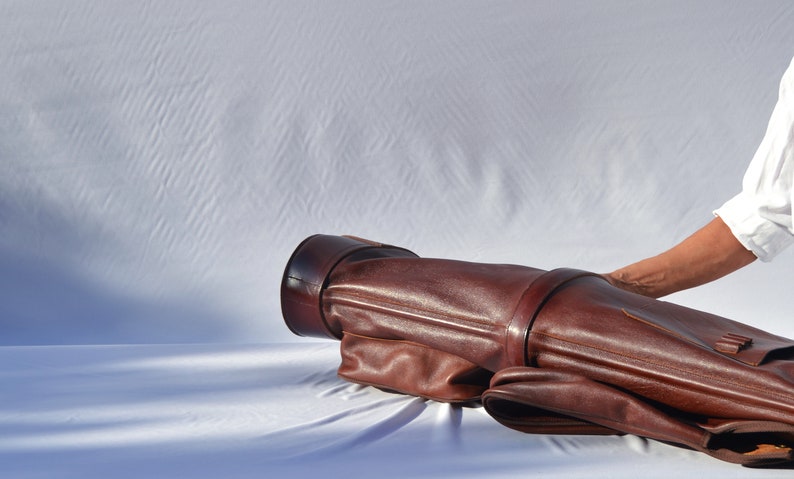 May include: A brown leather golf bag with a strap and buckle. The bag is lying on a white surface.