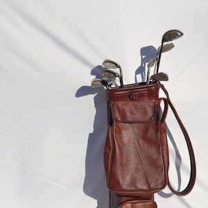 May include: A brown leather golf bag with a zipped pocket and a strap. The bag is filled with golf clubs.