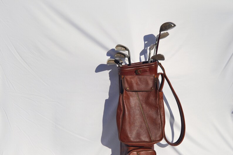 May include: A brown leather golf bag with a zipped pocket and a strap. The bag is filled with golf clubs.