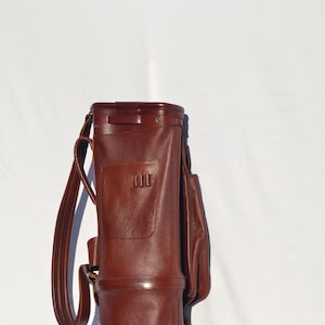 May include: A brown leather golf bag with a strap and a pocket. The bag is standing upright on a white surface.