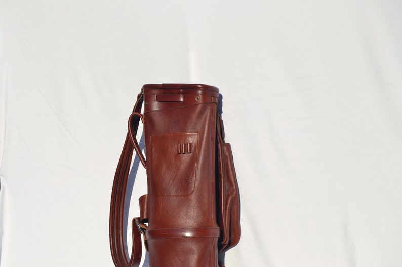 May include: A brown leather golf bag with a strap and a pocket. The bag is standing upright on a white surface.