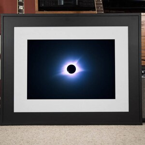May include: A framed photograph of a total solar eclipse. The sun is completely blocked by the moon, creating a dark circle surrounded by a bright halo of light.