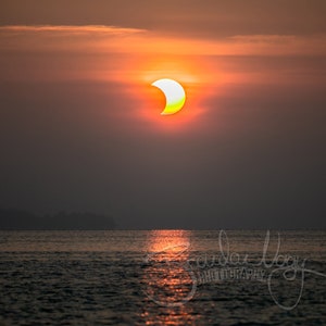Peut inclure: Une éclipse solaire partielle alors que le soleil se couche sur une étendue d'eau. Le soleil est d'un orange et d'un jaune vifs, avec un mince croissant de lune qui en bloque une partie. Le ciel est d'un orange et d'un rose doux, et l'eau est d'un bleu foncé.