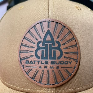 May include: A tan baseball cap with a brown leather patch. The patch features the text "BATTLE BUDDY ARMS" and a logo with bullets. The cap is made of a durable fabric.