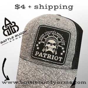 May include: Gray and black trucker hat with a patch featuring a skull, stars, and the words "PATRIOT" and "WHEN TYRANNY BECOMES REBELLION BECOMES DUTY." The hat has a mesh back and the website www.battlebuddyarms.com.
