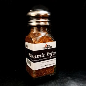 Może przedstawiać: Szklany słoik ze srebrną pokrywką wypełniony brązową solą morską infuzowaną balsamicznie. Etykieta zawiera napis "She Peppers Balsamic Infus A touch of savory to your dish with Sea salt infused with Contains: Aged Balsamic Vinegar and Sea Salt www.ShePeppers.com"