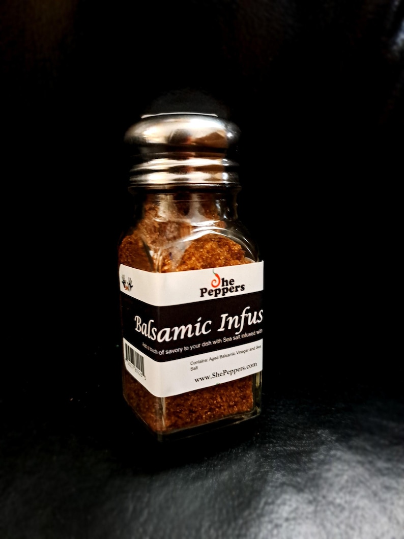 Może przedstawiać: Szklany słoik ze srebrną pokrywką wypełniony brązową solą morską infuzowaną balsamicznie. Etykieta zawiera napis "She Peppers Balsamic Infus A touch of savory to your dish with Sea salt infused with Contains: Aged Balsamic Vinegar and Sea Salt www.ShePeppers.com"